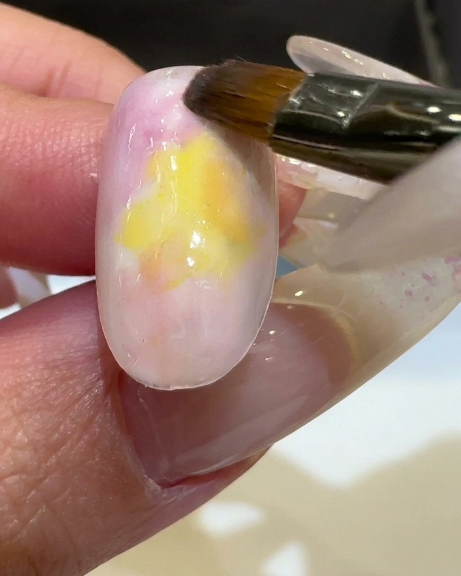 A brush applying a vibrant yellow gel polish to the center of a nail, blending into existing colors.