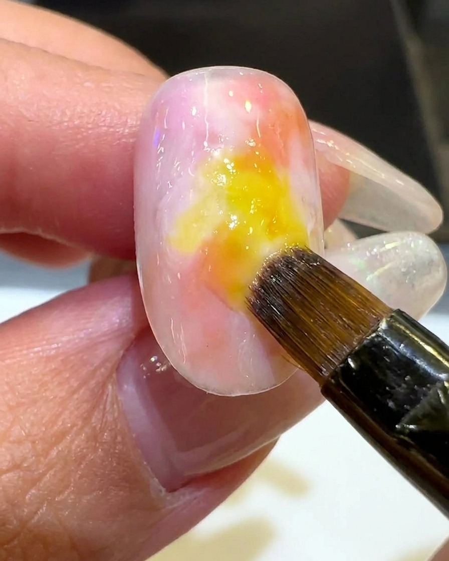 A brush applying a thin, sheer layer of milky white polish around the perimeter of the colored sunset gradient.