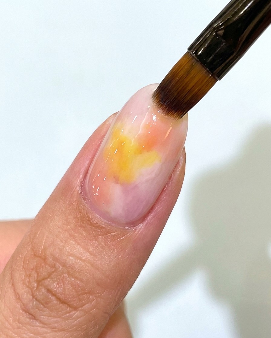 A nail brush applying a clear matte top coat over a pink and yellow sunset gradient gel nail.