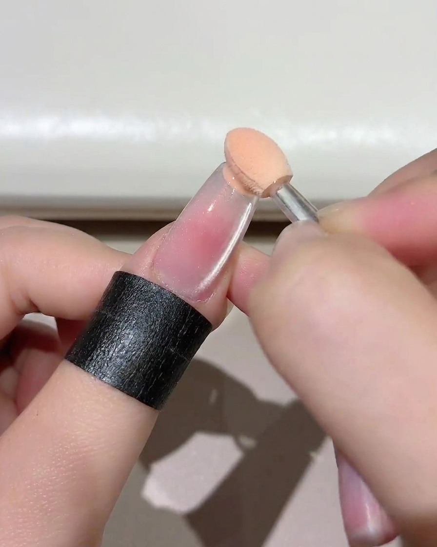 A close-up of a small sponge tool dabbing pinkish gel onto the free edge of a clear nail.