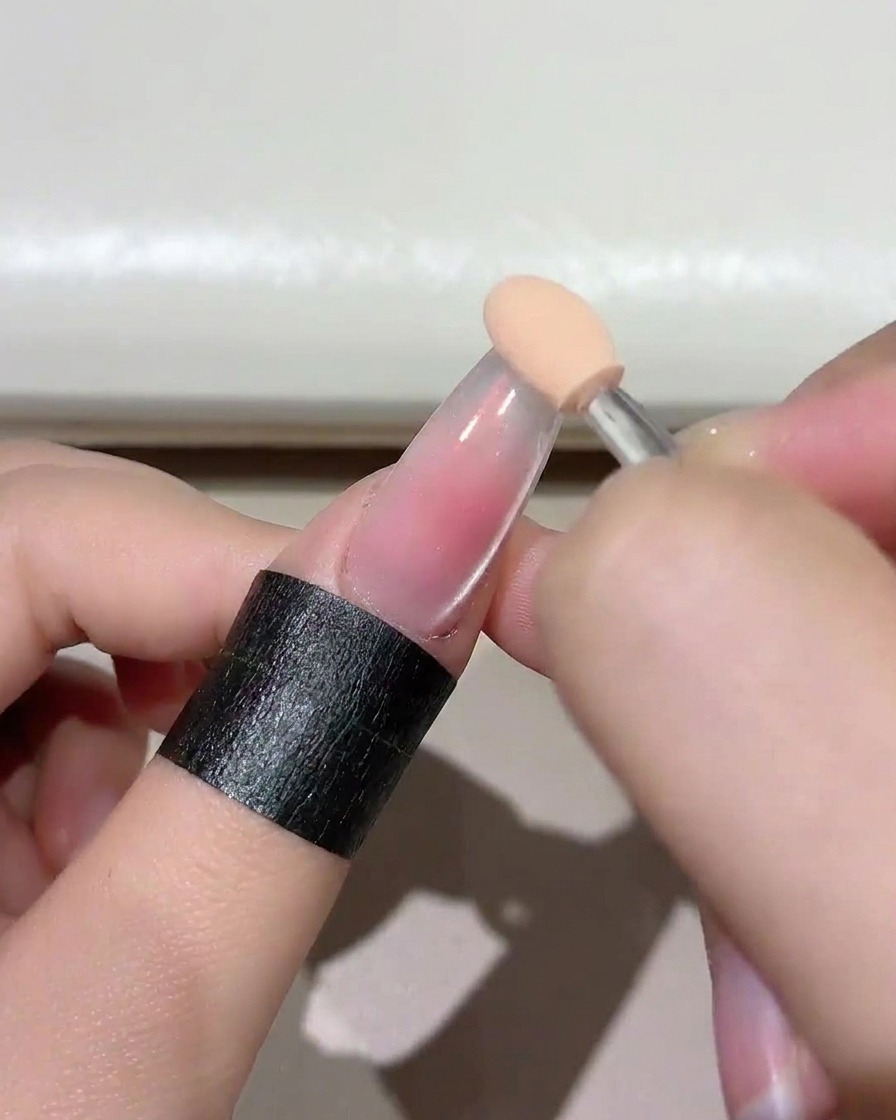 A close-up of a sponge applicator dabbing a soft blush pink gel onto the clear nail tip.