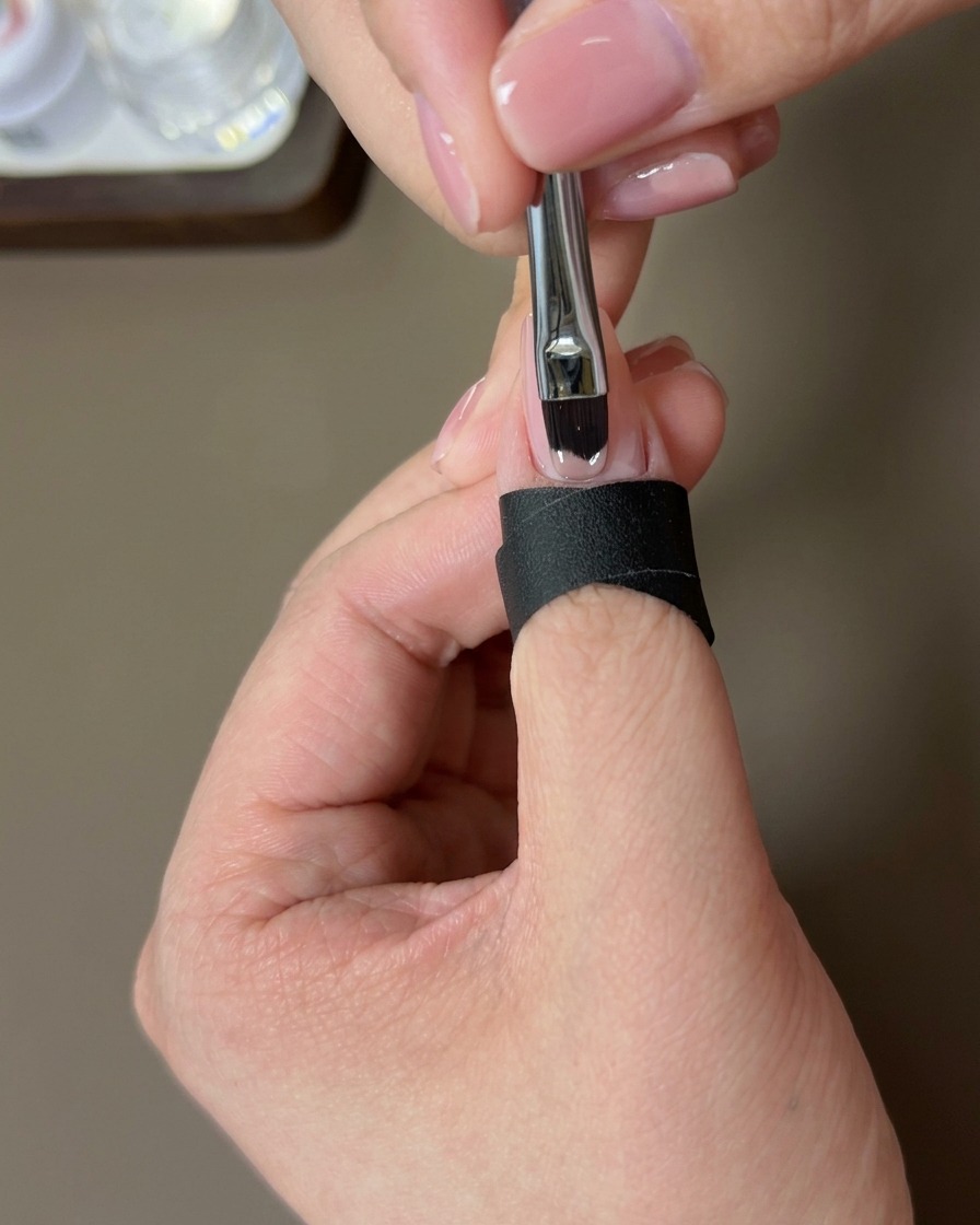 A brush applying a coat of nude gel polish to the natural nail.
