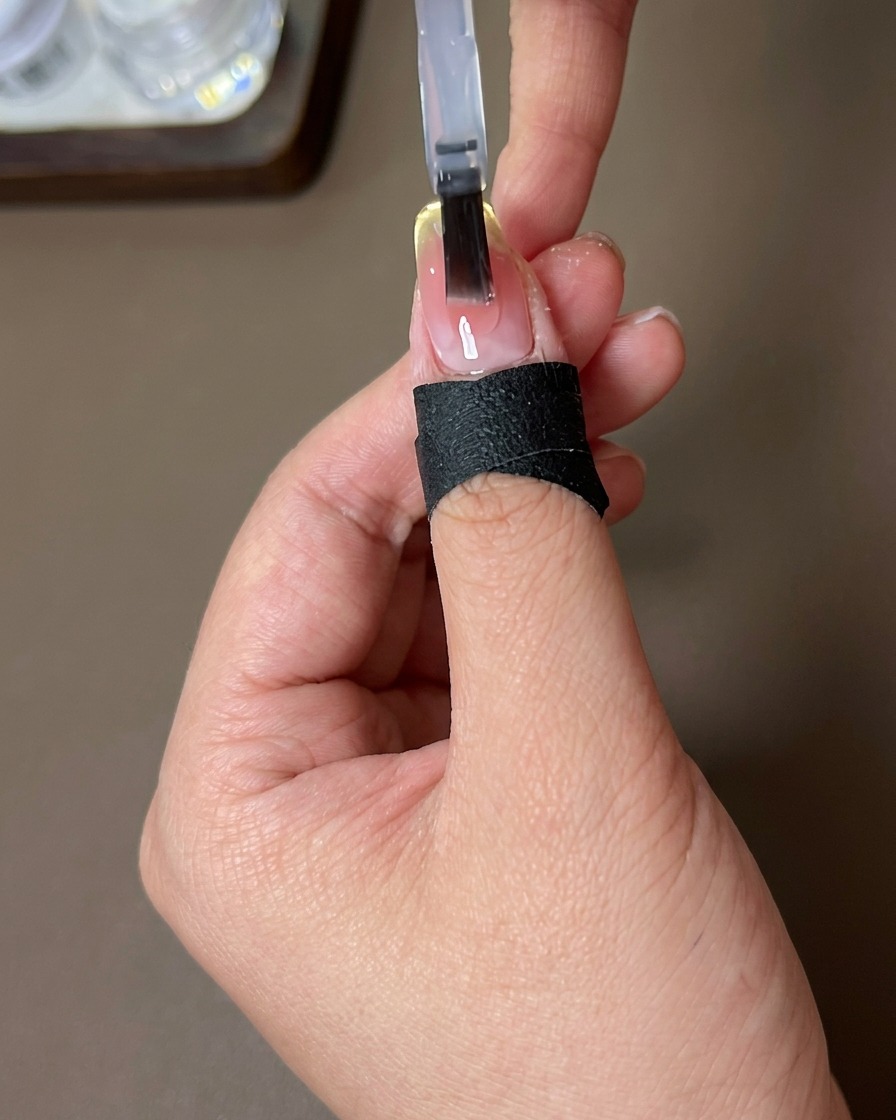 A brush applying a clear, glossy top coat layer onto a nail with a finished sunset-blush gradient and French-tip magnetic effect.