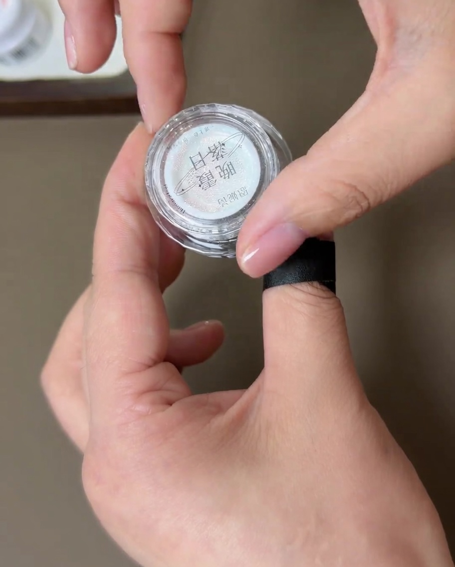 A small container of sunset aurora chrome powder being held with a hand that has a finished manicure, showing the powder texture.