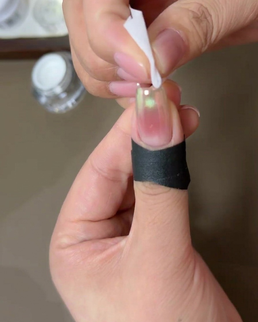 A person using a small lint-free wipe dampened with alcohol to clean the edges of a nail coated in aurora chrome powder.