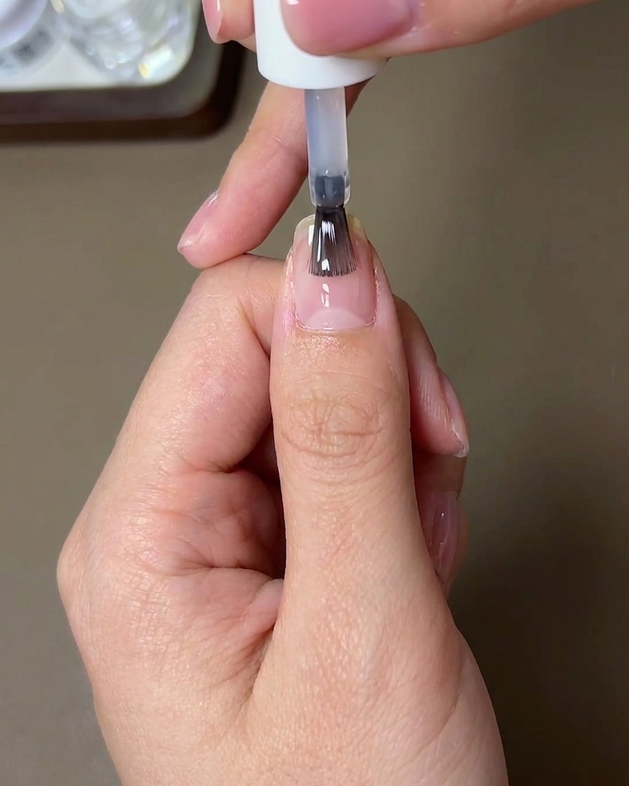 A brush applying a clear, glossy strengthening top coat over a nail finished with sunset aurora chrome powder.
