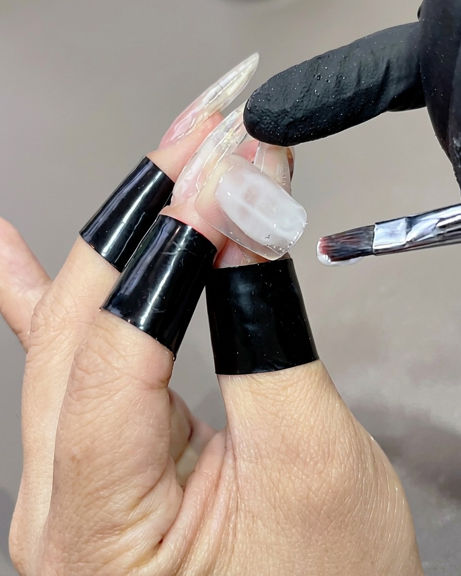A hand with long clear nail tips, where a flat brush is applying a milky white gel base coat to the ring fingernail.