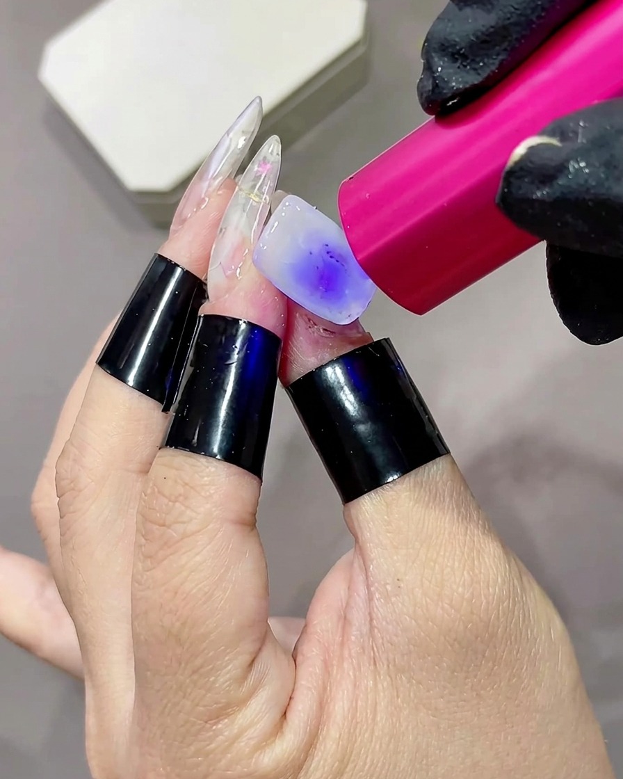 A close-up of a finger with a milky white base coat being cured by a small pink handheld UV or LED flashlight lamp.