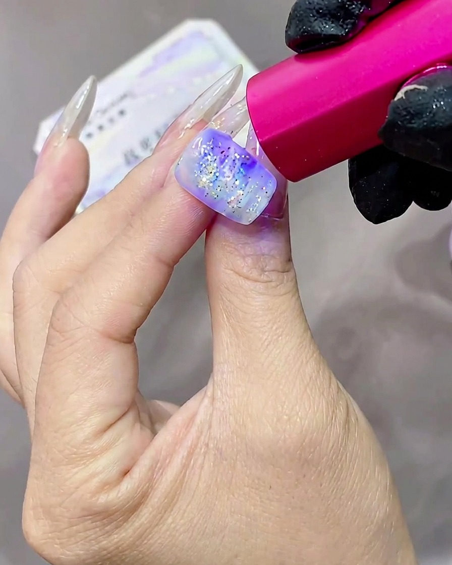 A hand with glittery, textured gel nails being cured under a pink-tinted UV lamp.