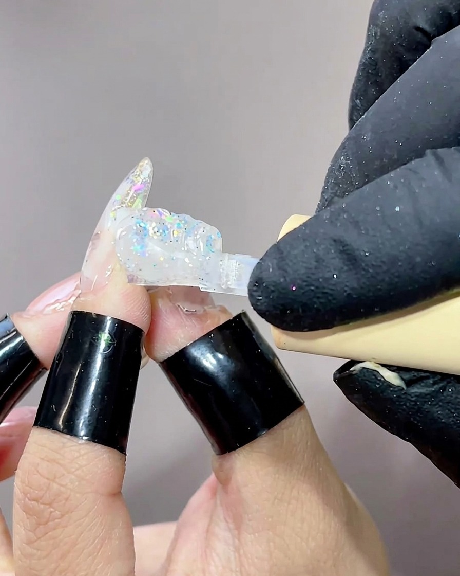 A close-up of a brush applying a clear, viscous builder gel to a nail already decorated with aurora-effect polish.