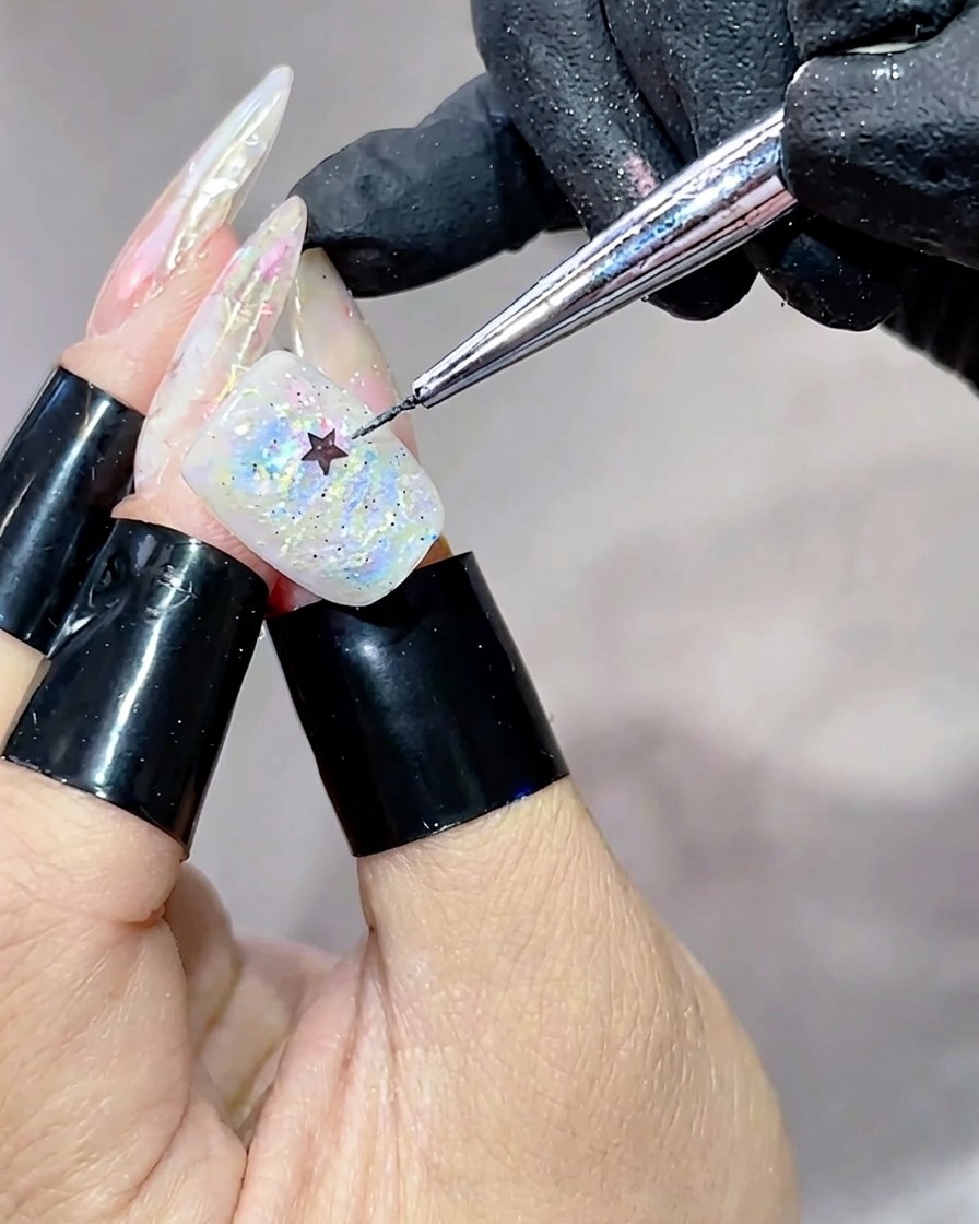 A metal tool placing a small black star-shaped sequin onto the center of a nail covered in iridescent, multi-colored gel.
