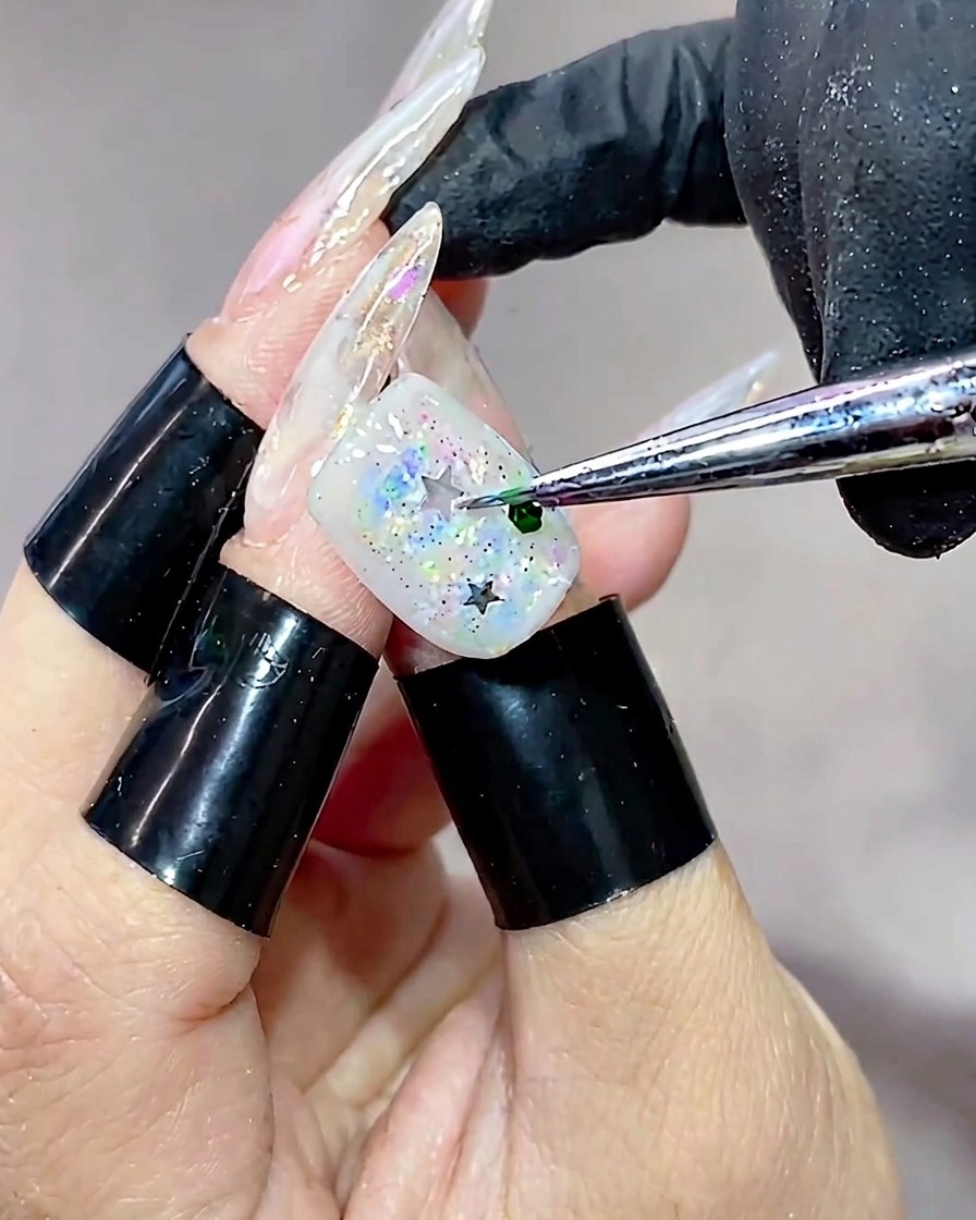 A close-up view of a tool placing a small, hexagonal green sequin onto a nail surface already adorned with star glitter.