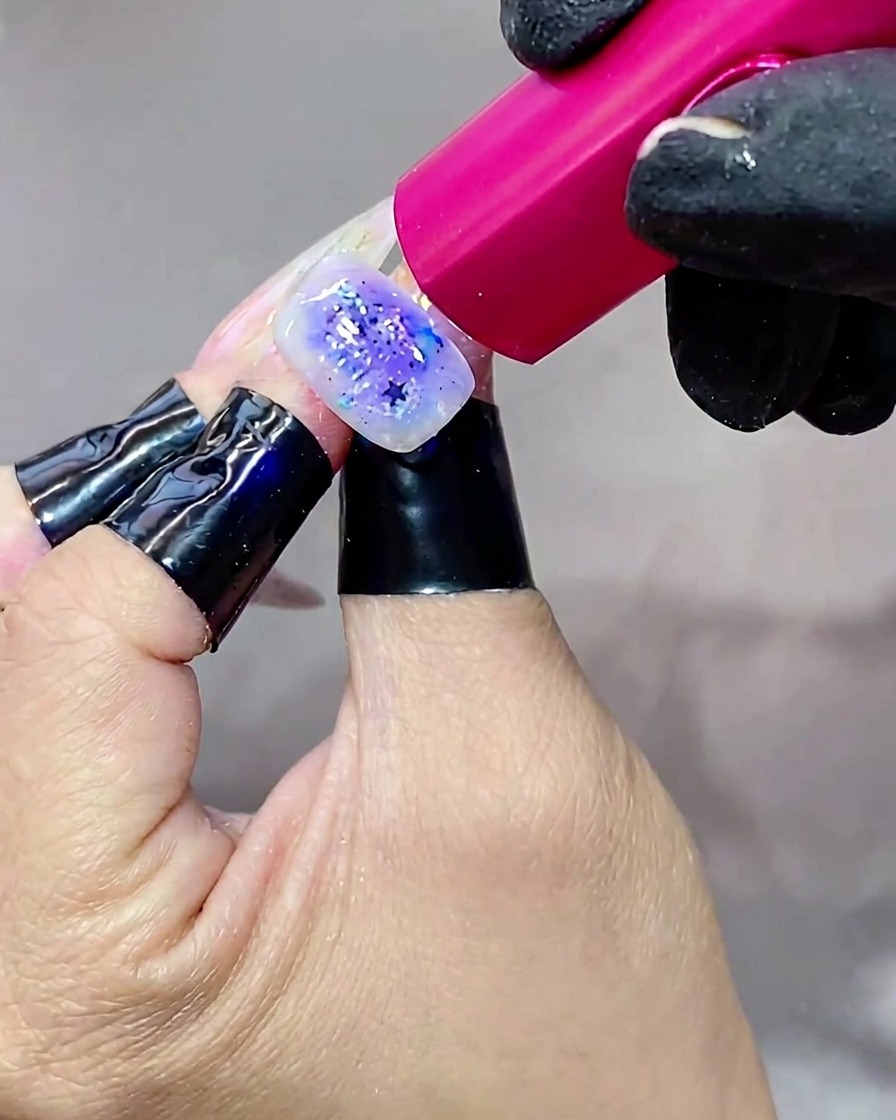 A hand holding a nail under a bright UV lamp, curing the final top coat layer.