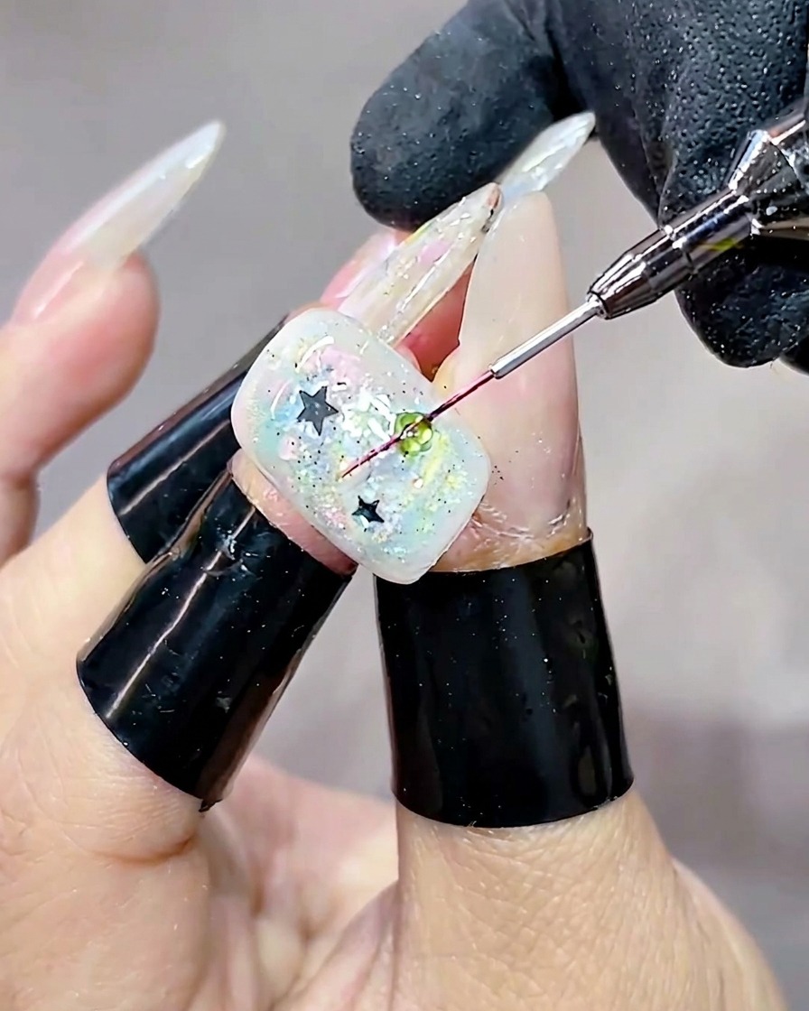 A close-up shot showing a fine brush applying a clear, rounded droplet of hard gel over a green sequin on a decorated nail.
