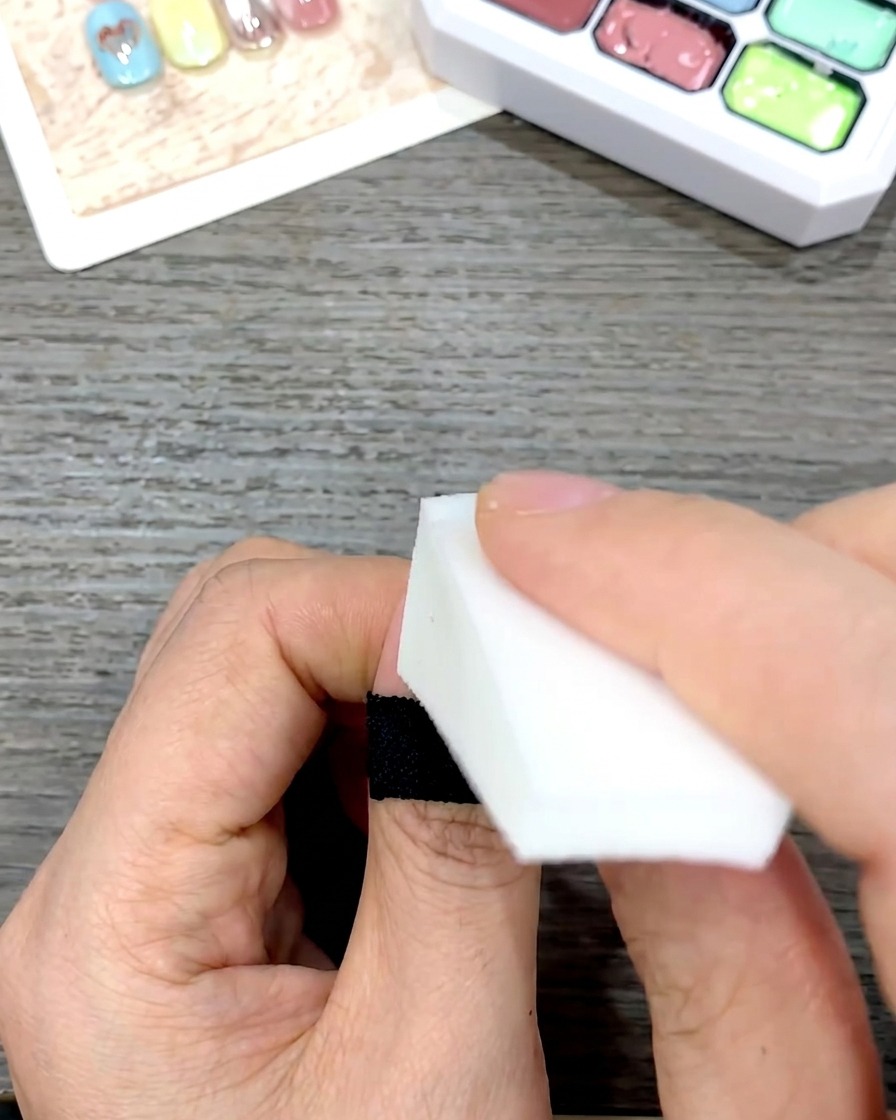 A hand using a white cosmetic wedge sponge to gently pat and blend yellow gel polish downward on a nail.