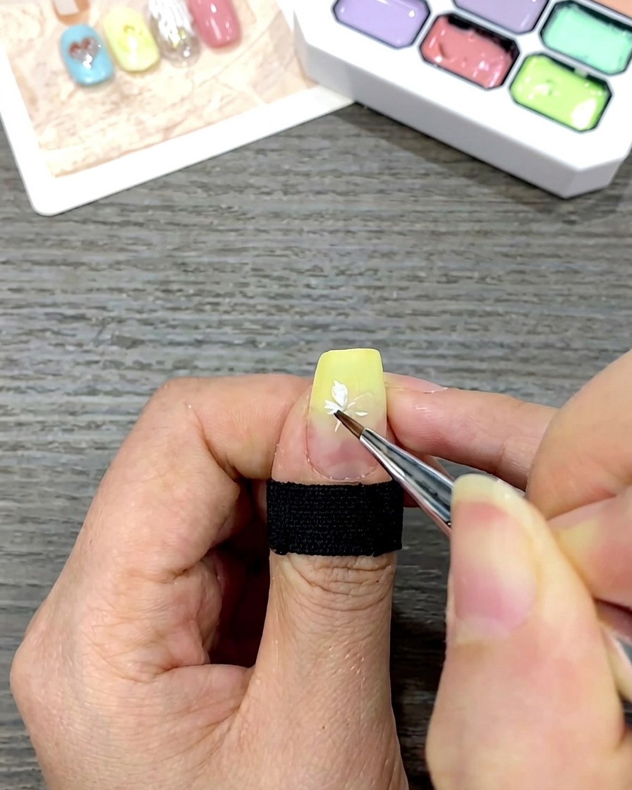 A close-up of a nail showing a completed white daisy flower painted on top of a yellow gradient base.