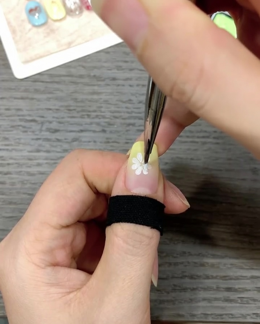 A close-up view of a nail being decorated with a fine liner brush applying a small, bright yellow dot to the center of a white daisy design.