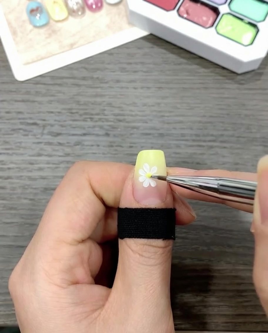 A brush applying a clear, glossy top coat over a nail decorated with a daisy design on a gradient background.