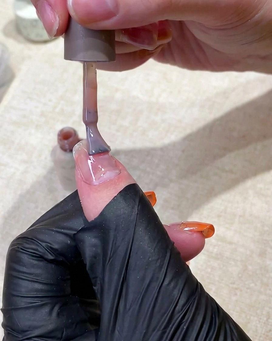 A hand in a black glove using a brush to apply a translucent nude gel base coat to a fingernail.