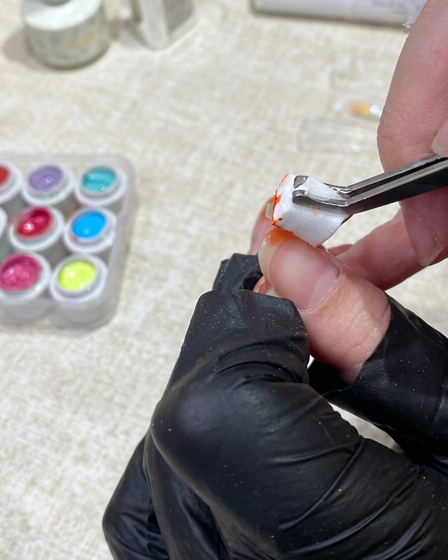 A gloved hand using tweezers to dab orange gel polish from a small sponge onto the tip of a fingernail.