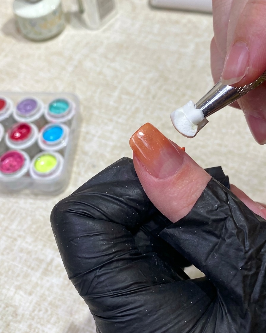 A tool holding a clean white sponge piece, gently tapping the middle of a fingernail to diffuse orange gel polish into a nude base.