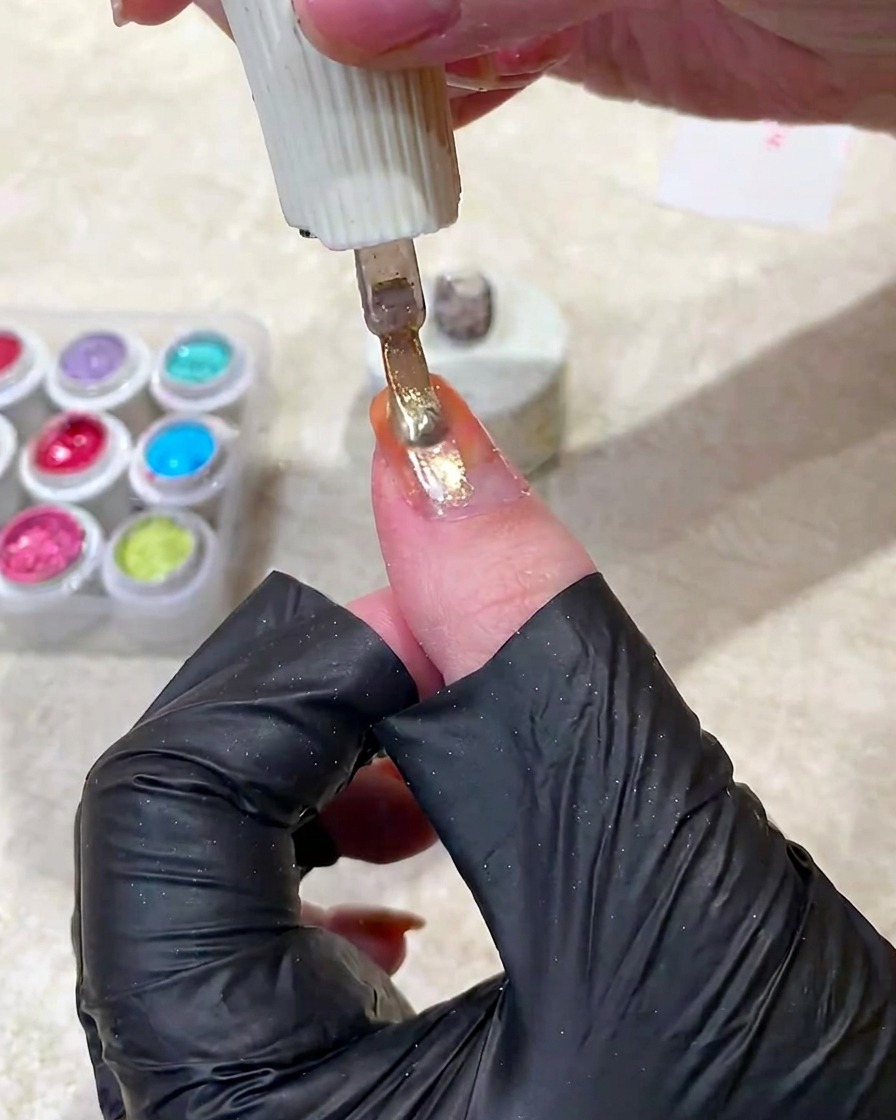 A nail brush applying a shimmering gold cat-eye gel polish over the orange gradient nail.
