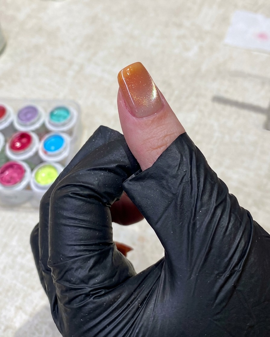 A hand wearing a black glove uses tweezers to hold a small sponge applicator, gently patting bright orange gel polish onto the tip of a manicured nail.