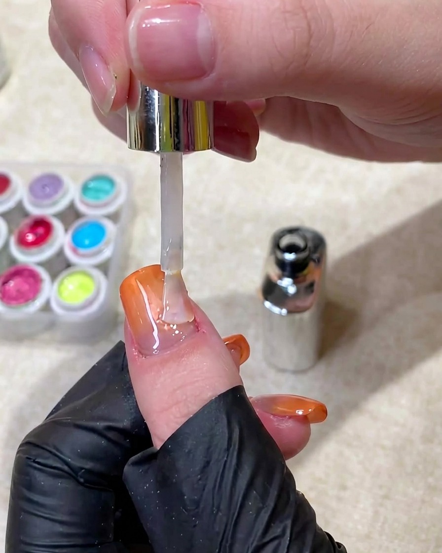 A brush applying a clear, glossy top coat over a finished orange gradient cat-eye nail.
