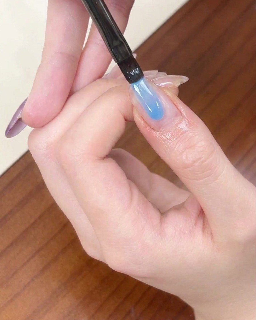 A liner brush applying a strip of sheer blue gel polish to the tip of an almond-shaped fingernail.
