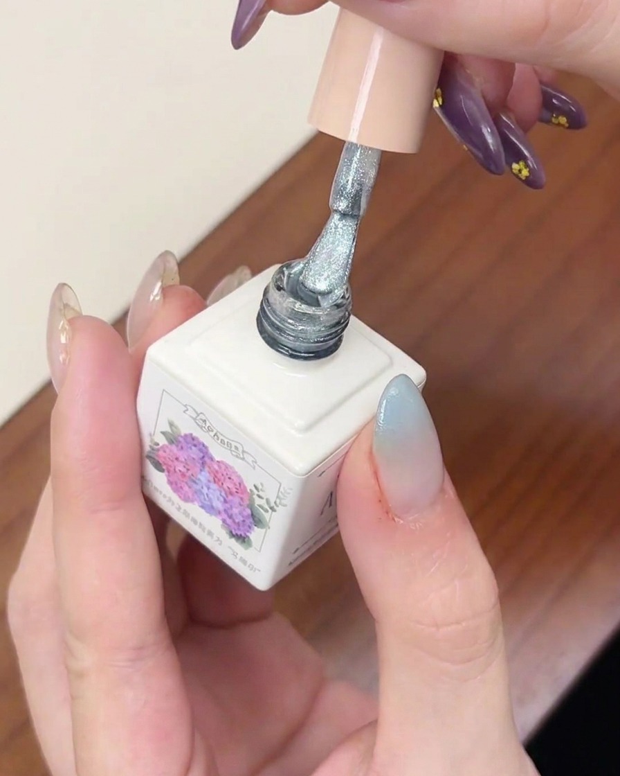 A gel polish brush applying a layer of silver shimmery cat-eye gel over a blue gradient fingernail.