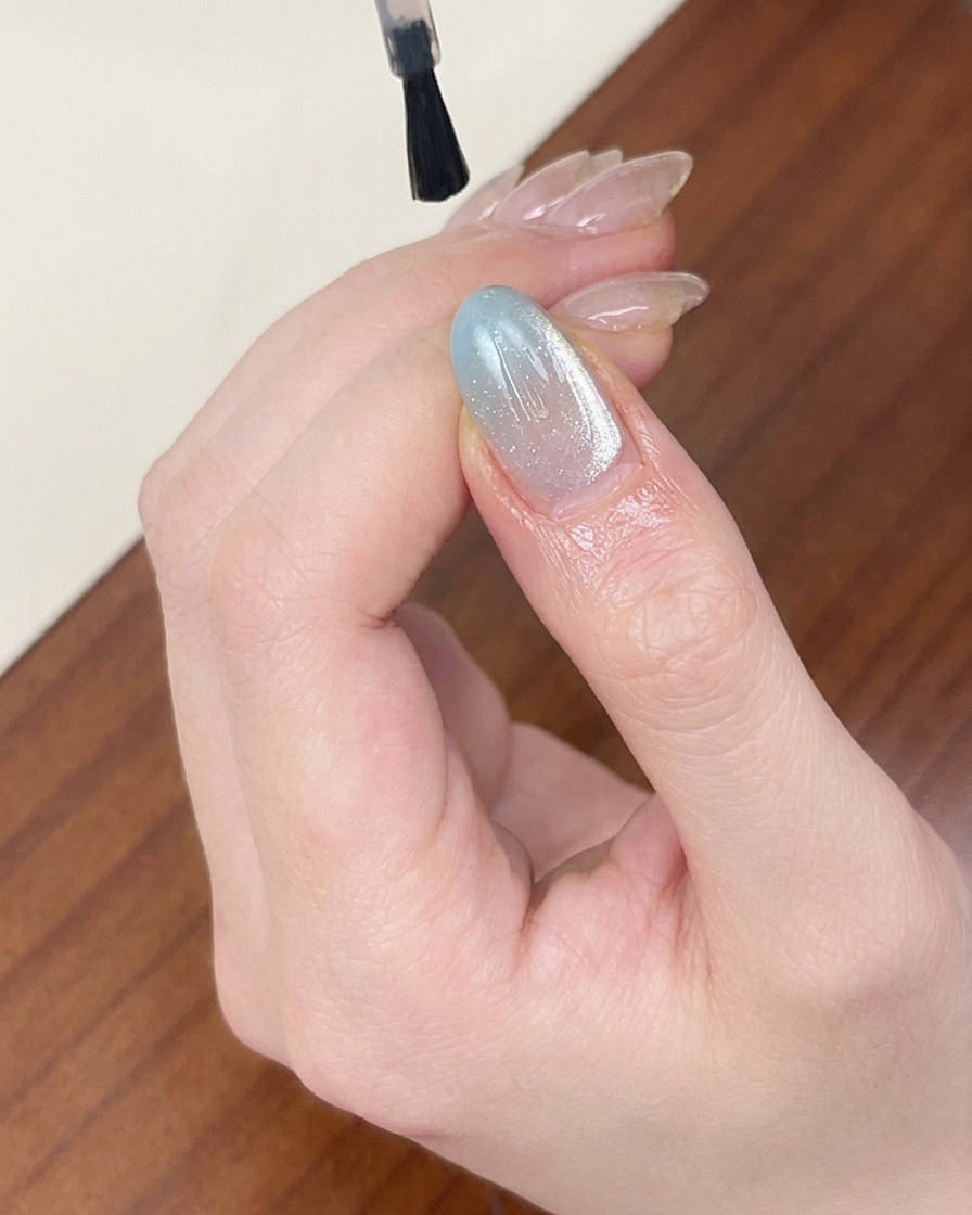 A brush applying a clear, glossy top coat over a light blue shimmery cat-eye gel nail.