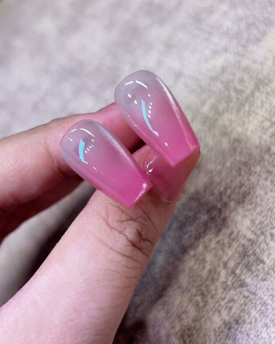 A brush applying a coat of shimmering white cat-eye gel polish over the existing pink ombre nail.
