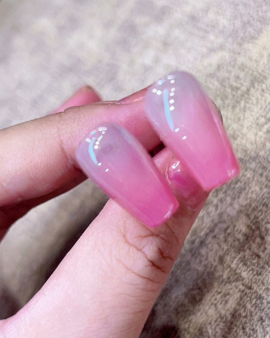 A close-up of a brush applying a clear, glossy top coat over a pink ombre nail.