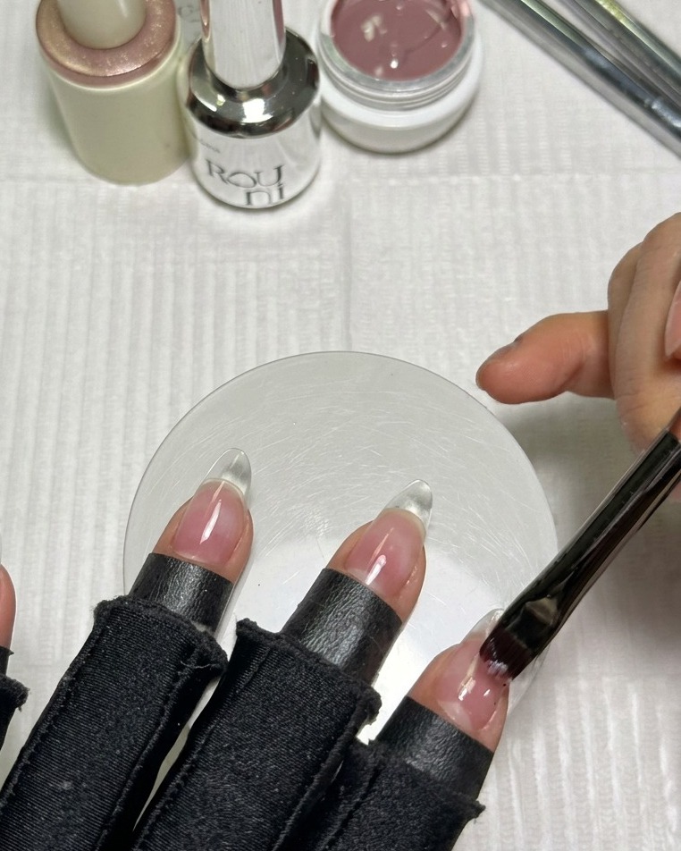 A brush applying a smooth layer of pinkish-nude gel polish onto a clear almond-shaped nail extension.
