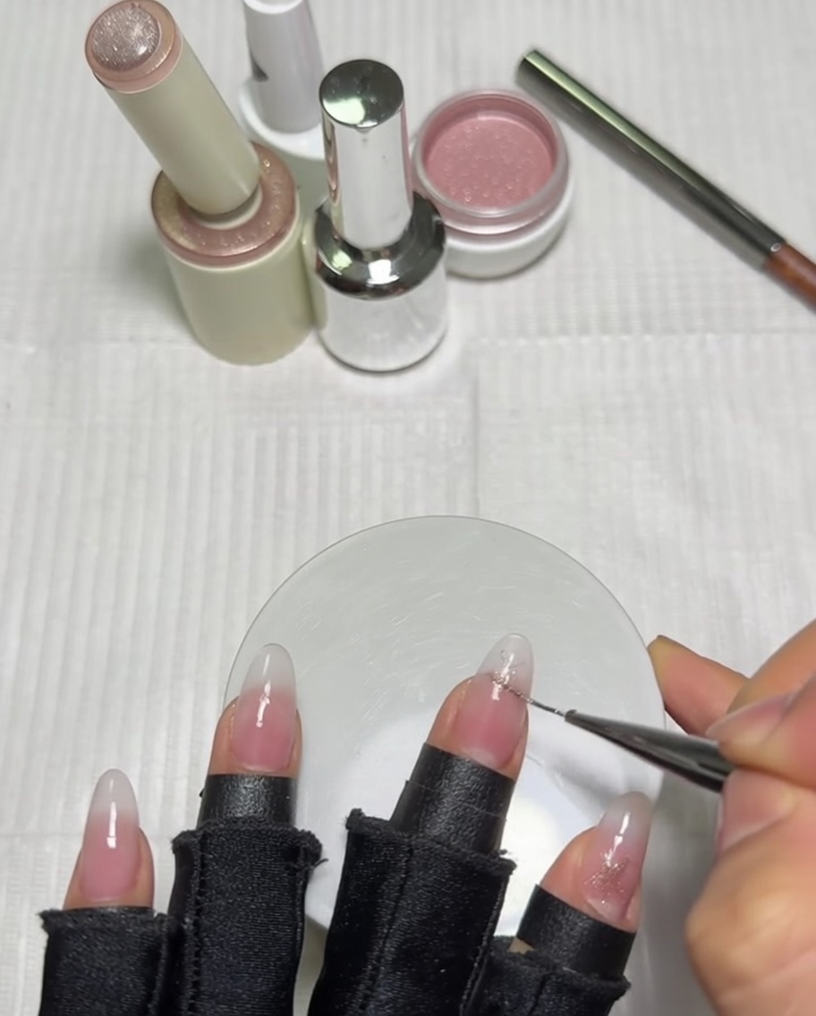 A close-up view of a hand using a long liner brush to detail a pink star design on a thumb nail that already has a milky gradient base.