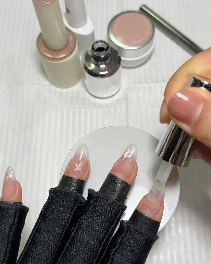 A brush applying a final glossy top coat over the completed star-decorated fingernails.