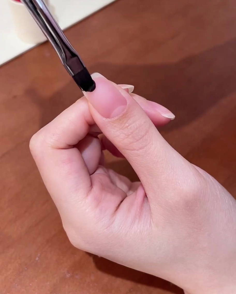 A close-up of a brush applying clear gel base coat to a clean natural nail.