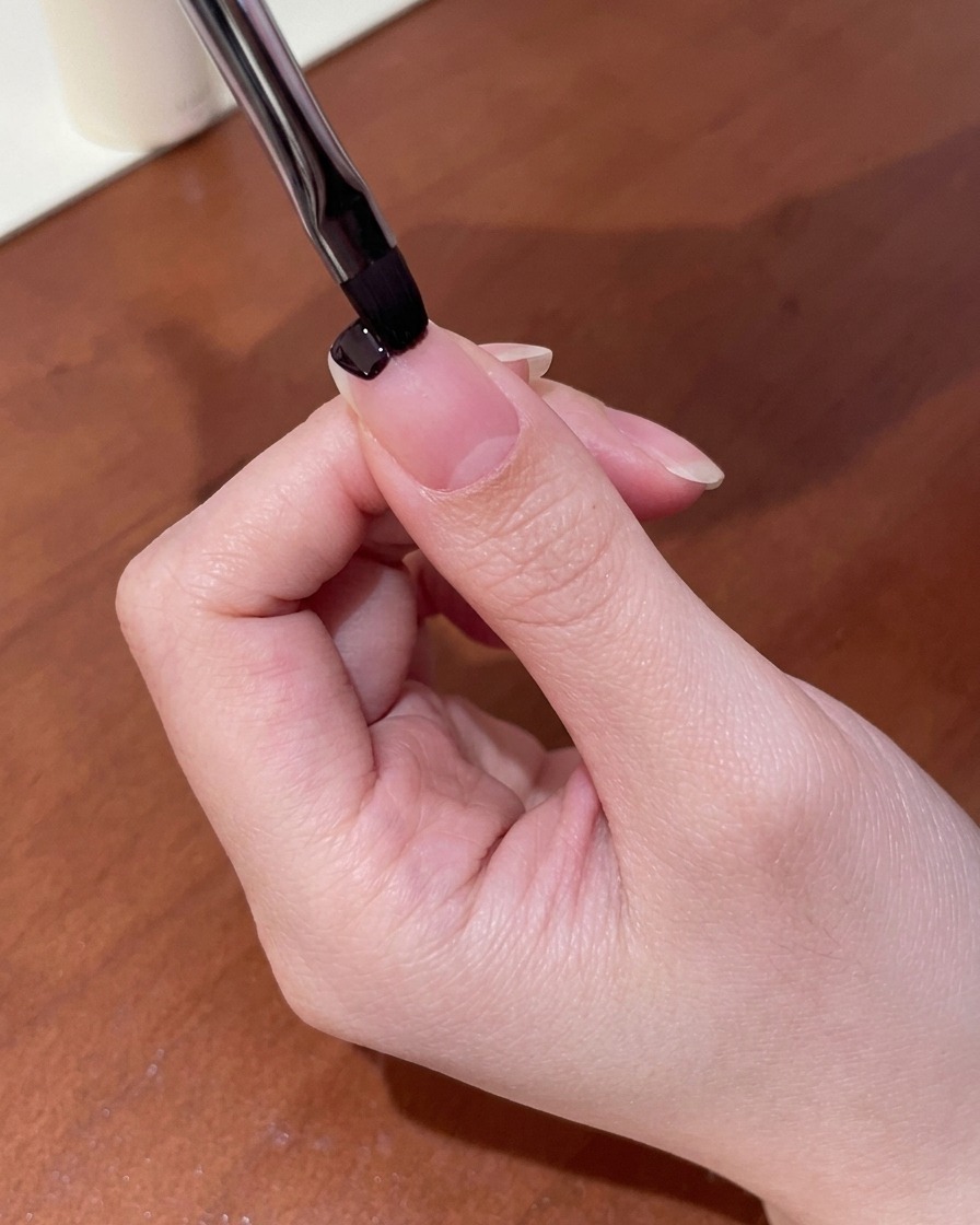 A close-up of a small nail brush applying dark purple gel polish to the tip of a fingernail.