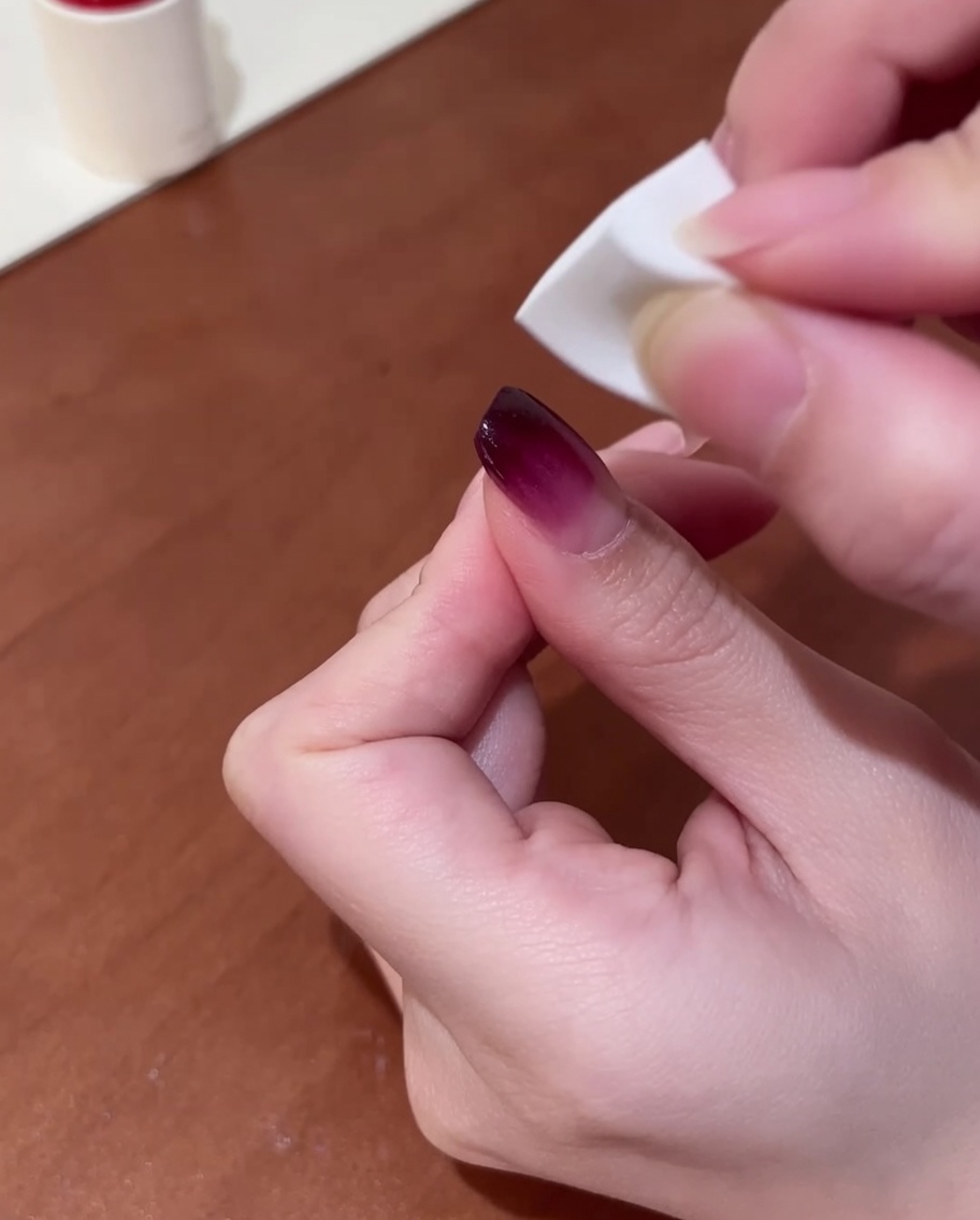 A close-up of a hand holding a small white makeup sponge, gently dabbing dark purple gel polish onto a fingernail to create a blended gradient finish.