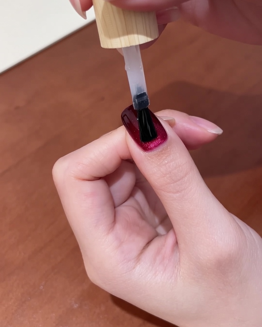 A close-up view of a hand with freshly applied top coat on dark red cat-eye nails, placed inside a UV nail lamp for the final curing step.
