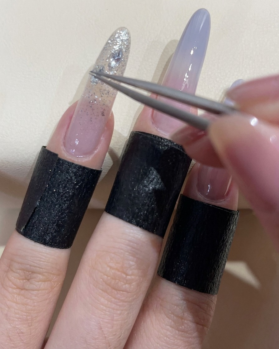 Tweezers placing a sparkling clear rhinestone onto a nail covered in wet adhesive gel.