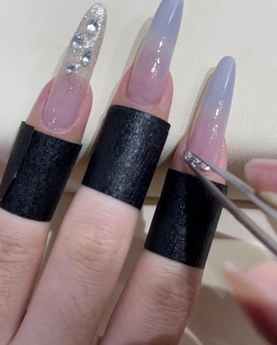 Close-up of tweezers applying small beads and stones around the cuticle of an index finger.