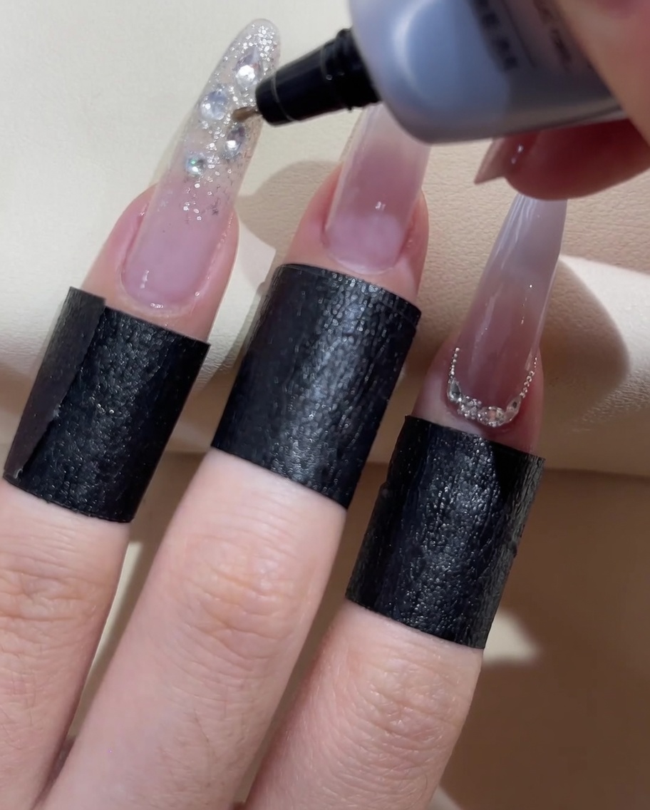 A fine-tip gel bottle applying clear sealant gel around the edges of decorative nail rhinestones.