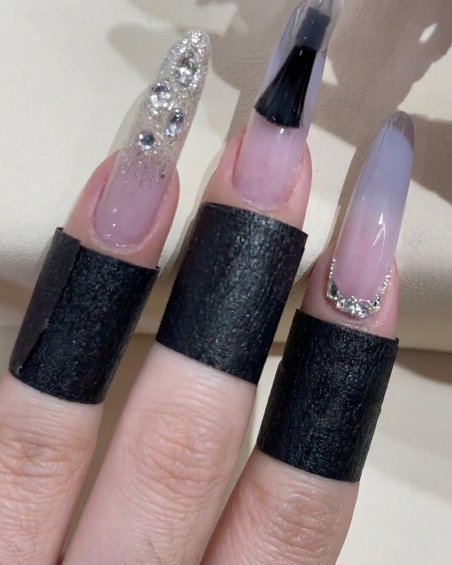 A close-up of a black nail polish brush actively applying a clear top coat to a light blue ombre nail, with adjacent finished fingers displaying silver glitter and rhinestone embellishments.