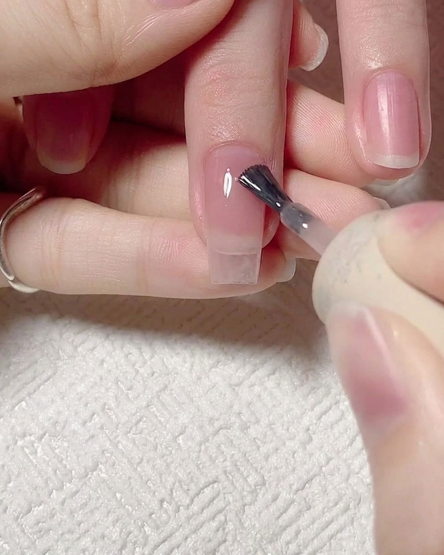 A close-up of a nail brush applying a sheer pink jelly gel to a clean, prepped natural nail.