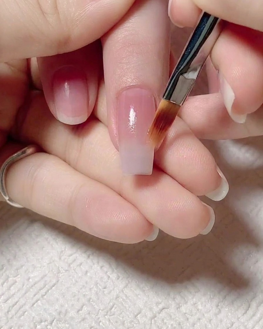 A flat brush gently pulling the jelly pink base coat upward on the nail to create a soft color gradient.