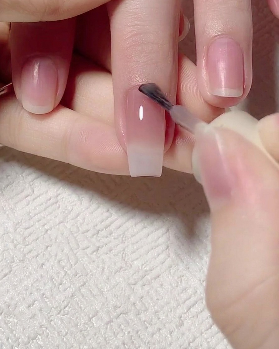 A brush applying a jade-tinted pink gel to the cuticle area of the nail.