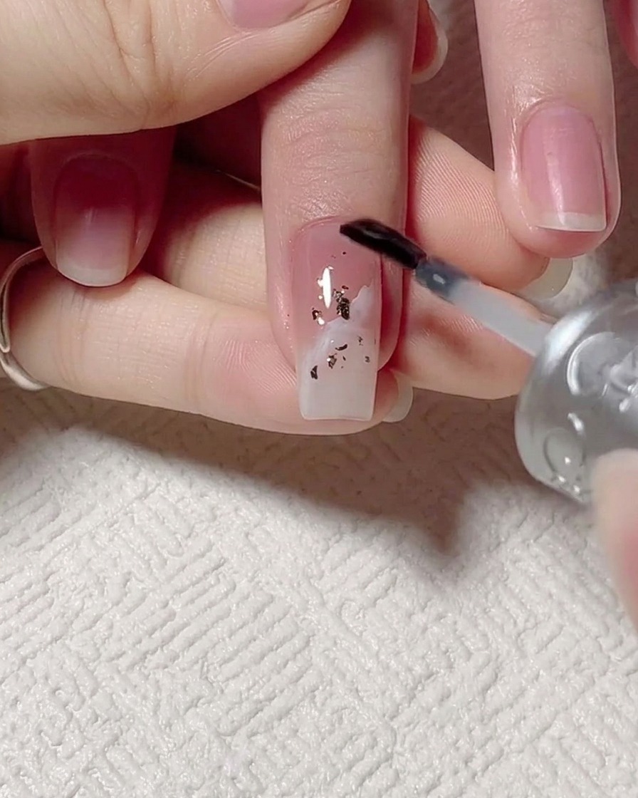 A brush applying clear base gel over a nail with gold foil and wave designs.