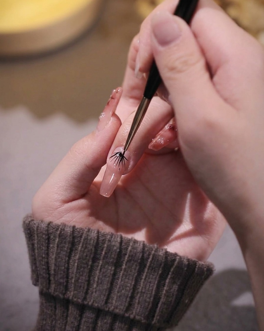 A close-up of a fine nail art liner brush refining the edges of pink gel on a fingernail.