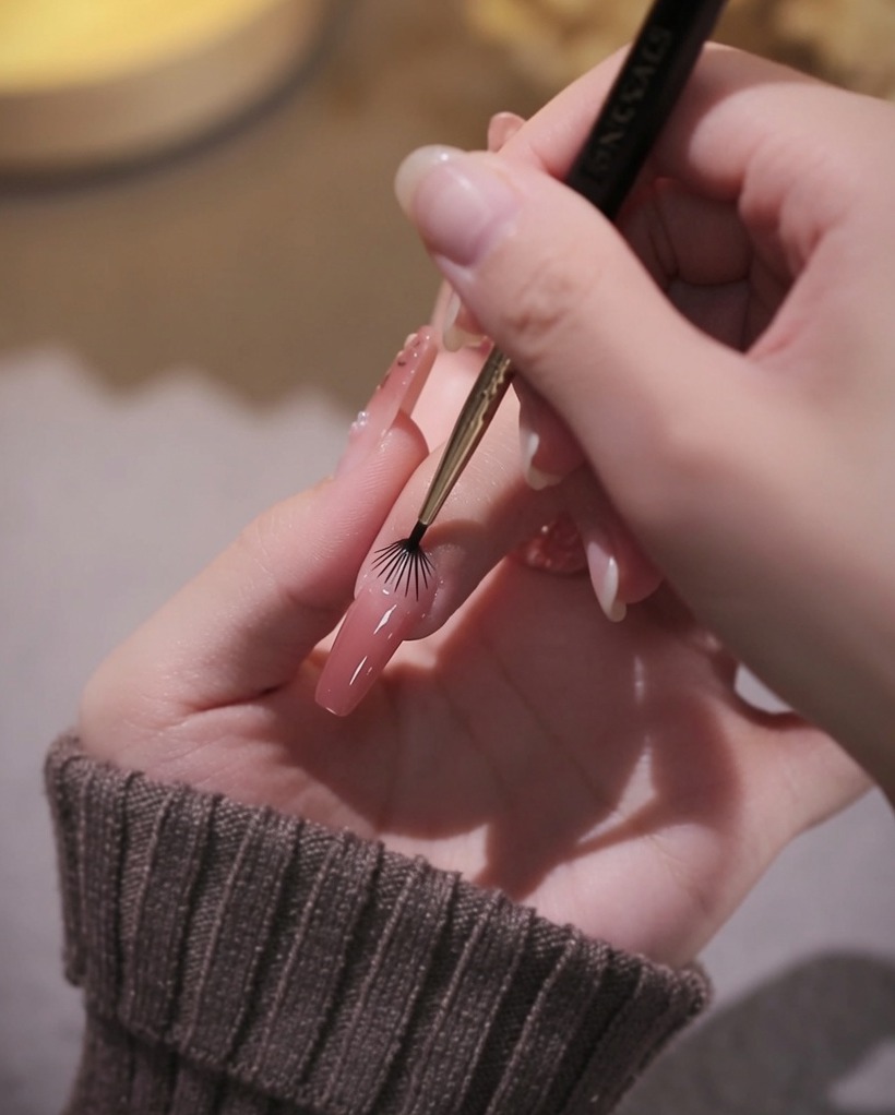 A close-up of a liner brush being used to carefully outline the cuticle area with pink gel polish.